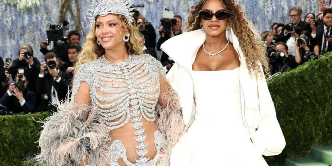 Two glamorous women pose on a red carpet at a Met Gala-like event, surrounded by photographers; one wears a crystal bodysuit and headdress, the other in a white outfit with sunglasses and pearls.