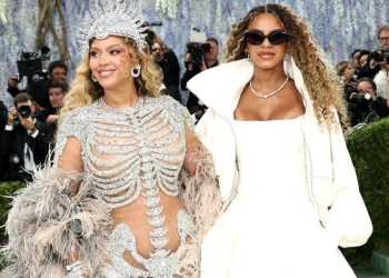 Two glamorous women pose on a red carpet at a Met Gala-like event, surrounded by photographers; one wears a crystal bodysuit and headdress, the other in a white outfit with sunglasses and pearls.