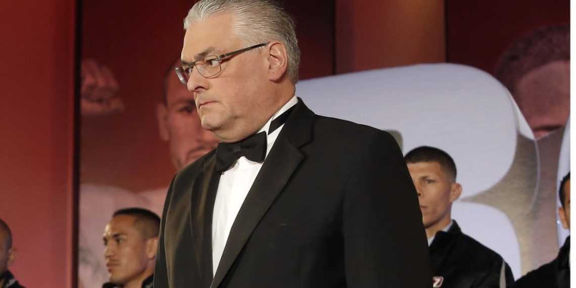 Man in a black tuxedo and bow tie on stage, looking to the left, with a blurred audience behind him.