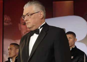 Man in a black tuxedo and bow tie on stage, looking to the left, with a blurred audience behind him.
