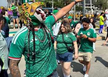 Crowd at an outdoor festival; a performer wearing a bright dragon mask and green patterned shirt leads a group in matching green jerseys.
