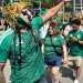 Crowd at an outdoor festival; a performer wearing a bright dragon mask and green patterned shirt leads a group in matching green jerseys.