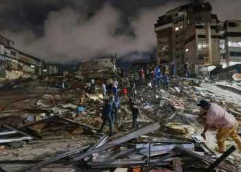 Nighttime rescue scene in a collapsed city block: rubble, damaged buildings, and people searching for survivors.