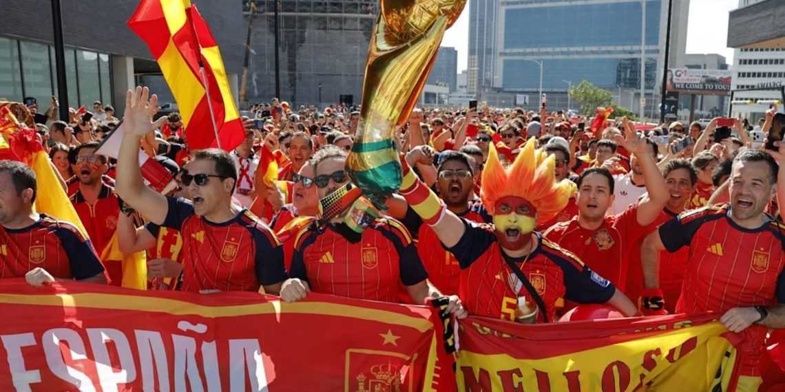 Crowd of Spain soccer fans in red and yellow jerseys, waving flags and cheering behind a red banner on a city street festival.