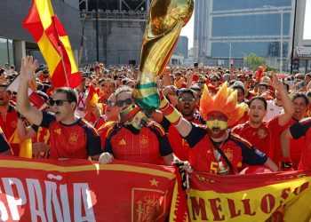Crowd of Spain soccer fans in red and yellow jerseys, waving flags and cheering behind a red banner on a city street festival.