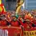 Crowd of Spain soccer fans in red and yellow jerseys, waving flags and cheering behind a red banner on a city street festival.