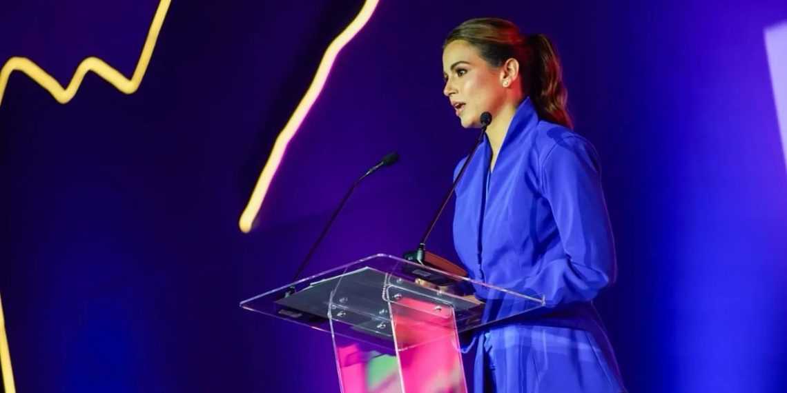 Woman in a blue suit delivering a speech at a clear acrylic podium on a purple-lit stage.