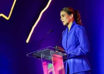 Woman in a blue suit delivering a speech at a clear acrylic podium on a purple-lit stage.