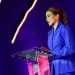 Woman in a blue suit delivering a speech at a clear acrylic podium on a purple-lit stage.