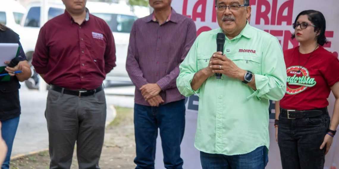 Man in a light green Morena shirt speaks into a handheld microphone at an outdoor event, with three people standing nearby behind him.