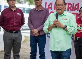 Man in a light green Morena shirt speaks into a handheld microphone at an outdoor event, with three people standing nearby behind him.