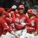 Red-uniform baseball players celebrate together on the field, with one central player raising a fist in triumph as teammates join in cheers.