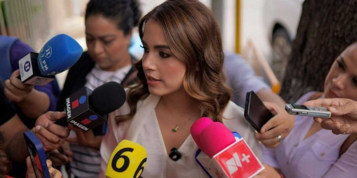 Woman being interviewed by several reporters with colorful microphones surrounding her outdoors.
