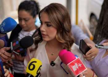 Woman being interviewed by several reporters with colorful microphones surrounding her outdoors.